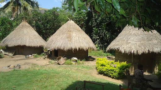 Three thatched-roof huts surrounded by lush greenery and a sunny garden with a seated person in the foreground.
