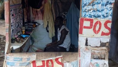 Man sitting in a small makeshift barbershop adorned with haircut posters and "POS" signs.