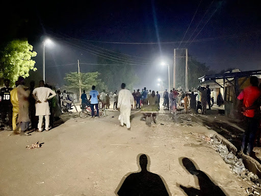 A nighttime street scene with a crowd of people gathered under streetlights, surrounded by trees and shadows.