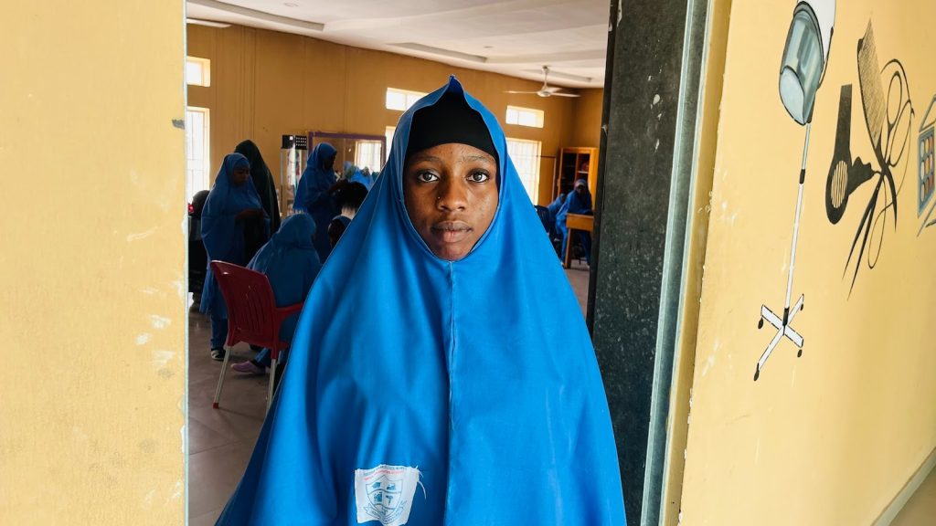 Young person in a blue hijab stands in a room with others in similar attire, with educational wall art visible.