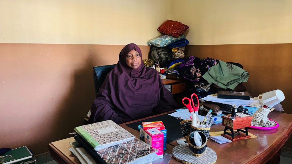 Person in a purple outfit sits at a cluttered desk in an office with various papers, fabric, and office supplies.