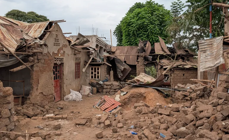 Collapsed buildings with debris in front, showing damage against a backdrop of trees and a cloudy sky.