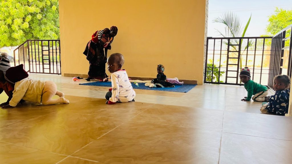 Babies crawl and sit on a tiled floor while a caregiver attends to one on a mat, surrounded by a calm, open environment.