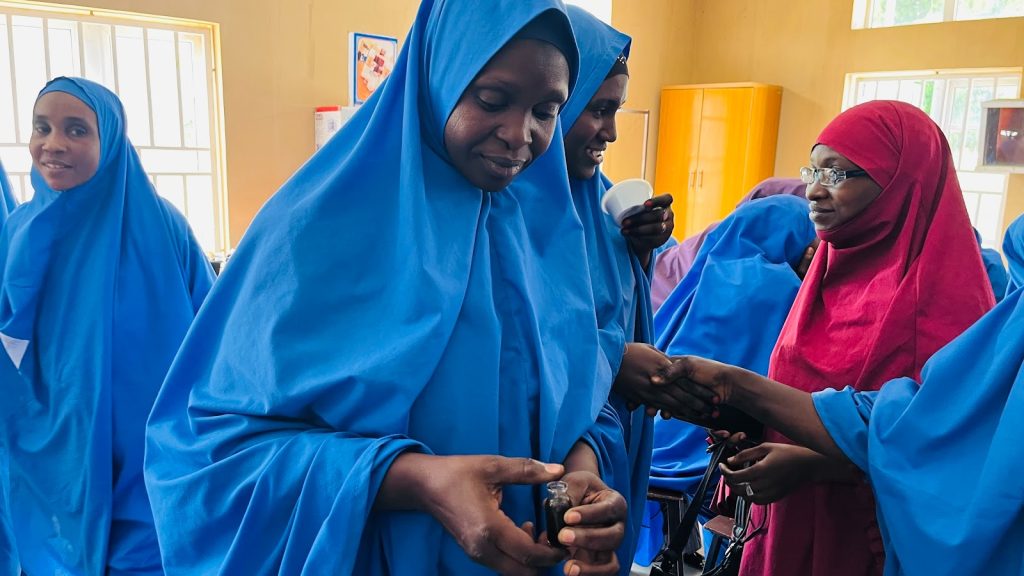 Group of women in blue and red hijabs gather, some holding bottles and mugs, in a brightly lit room.