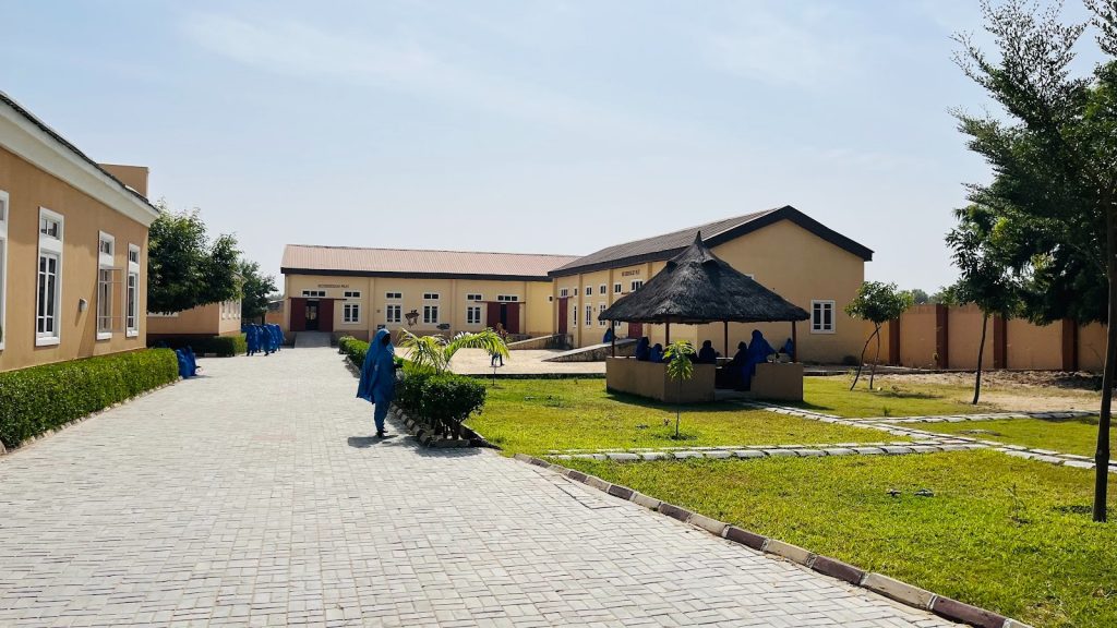 People in blue attire walk and sit in a courtyard with buildings and greenery under a clear sky.