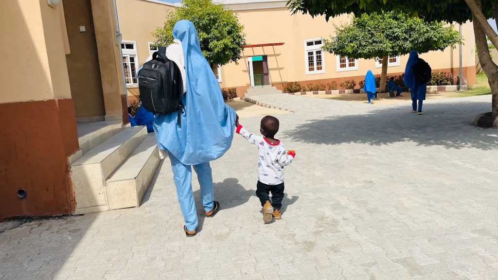 Child holding hands with a person in a blue outfit, walking along a sunny path near a building with trees.