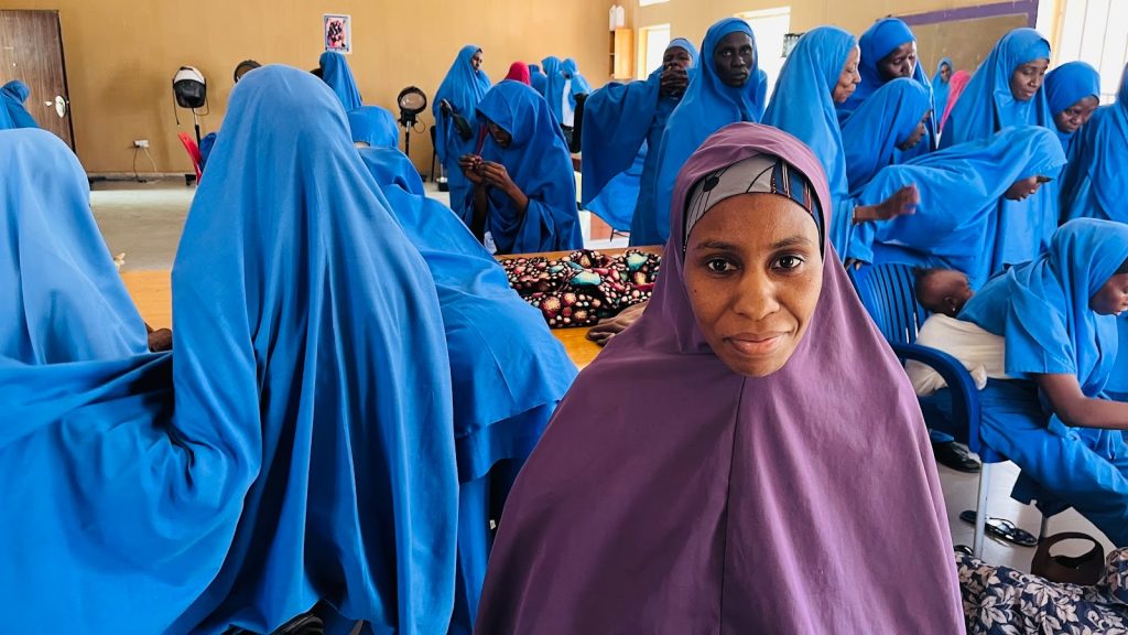 A group of women in blue garments gather around a table, with one woman in a purple garment smiling at the camera.