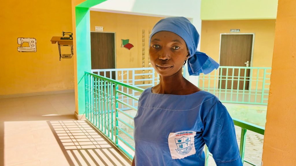 A woman in blue attire and headscarf stands in a sunny corridor with colorful walls and sewing machine mural.