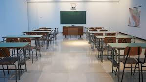 Empty classroom with rows of desks, chairs, a teacher's desk, and a blackboard. Bright lighting and a map on the wall.