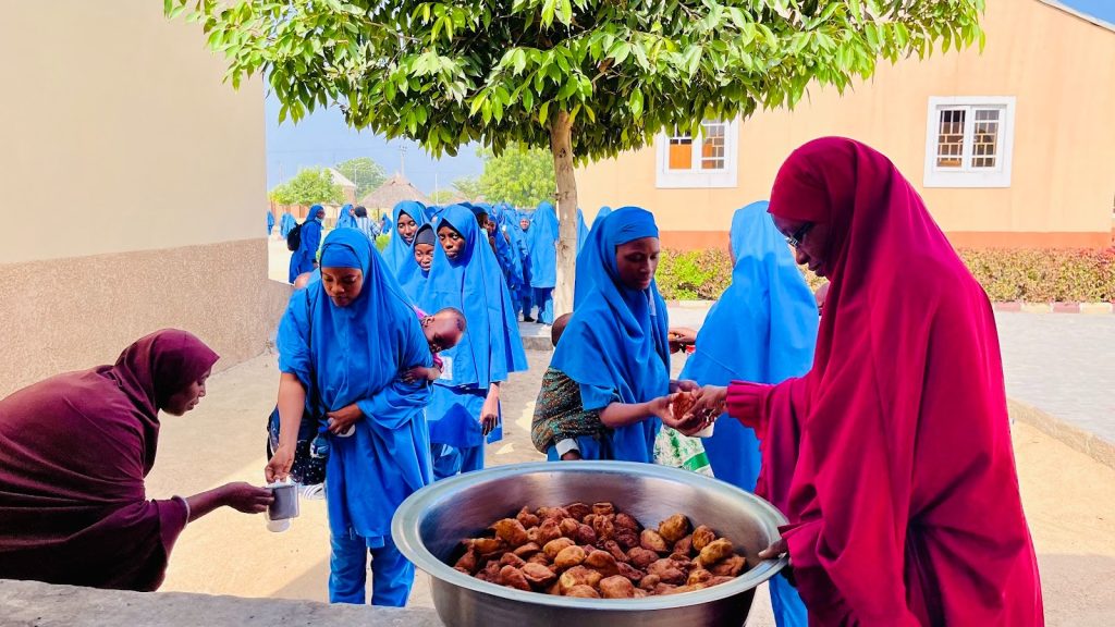 Women in blue uniforms queue for food from a large bowl outside, with one woman serving from a pot under a tree.