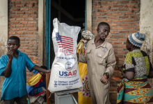 A man distributes a large bag of USAID food aid to two women while a boy stands nearby against a brick wall.