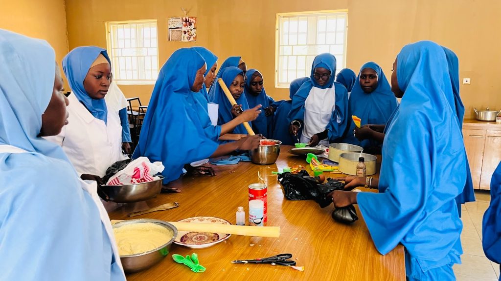 Group of students in blue uniforms engaged in a baking class, gathered around a table with mixing bowls and ingredients.