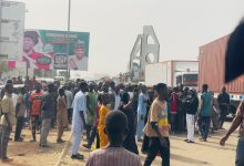 A large crowd gathers near trucks and a billboard on a busy street with an arch in the background.