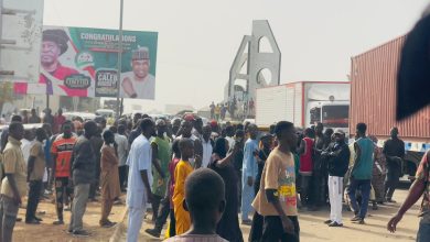 A large crowd gathers near trucks and a billboard on a busy street with an arch in the background.