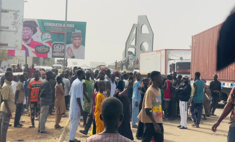 A large crowd gathers near trucks and a billboard on a busy street with an arch in the background.