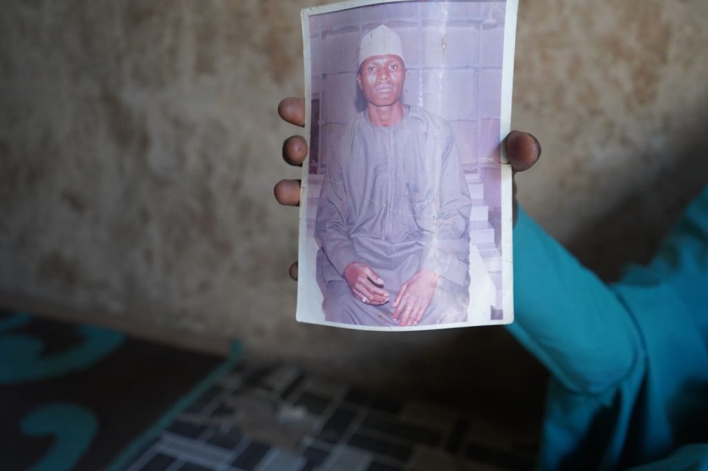 A hand holding a photograph of a man sitting and wearing a traditional outfit and cap.