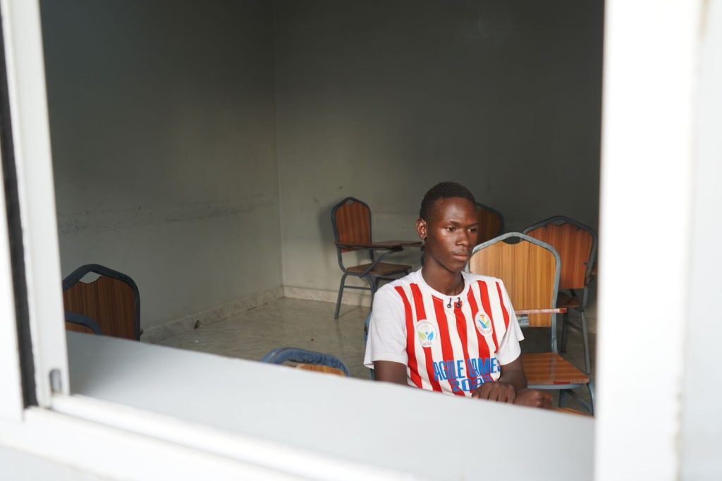 Young man in a red and white striped shirt sits alone in a sparse room with empty chairs.