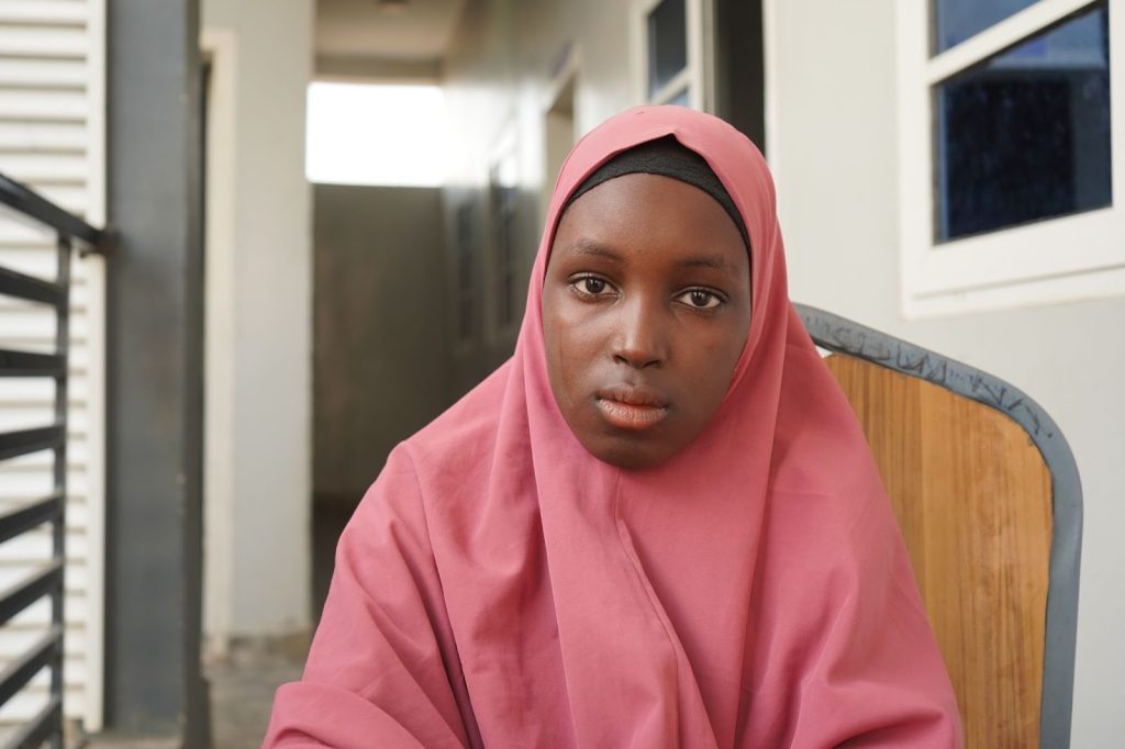 A person wearing a pink hijab sits on a wooden chair in a corridor, looking at the camera.