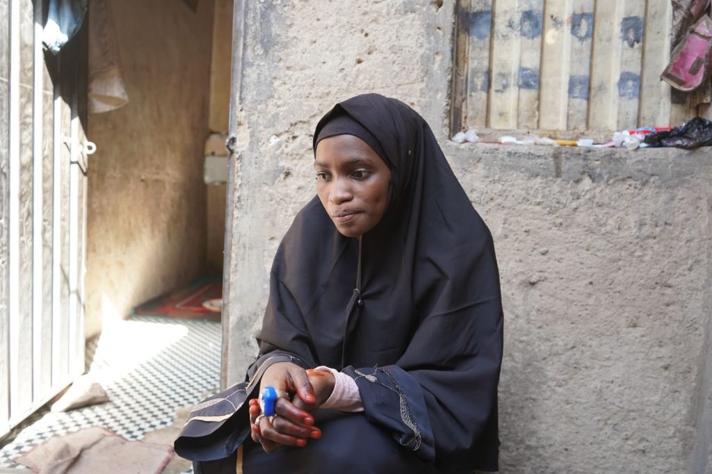 A woman in a black hijab sits pensively outside a building, holding a blue object in her hand.