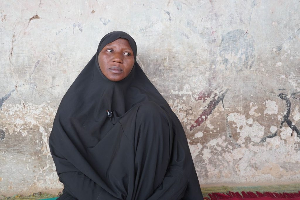 Woman in a black headscarf and robe sitting against a textured wall, looking pensive.