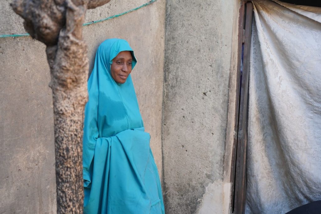 Woman in a turquoise hijab stands against a concrete wall outdoors, next to a tree and a doorway with a white curtain.