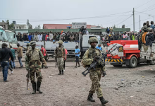 Soldiers patrol an area with trucks and people gathered in the background in a busy outdoor setting.