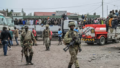Soldiers patrol an area with trucks and people gathered in the background in a busy outdoor setting.