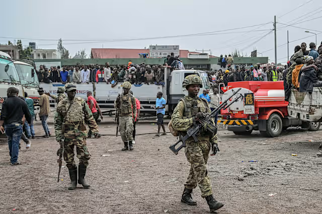 Soldiers patrol an area with trucks and people gathered in the background in a busy outdoor setting.