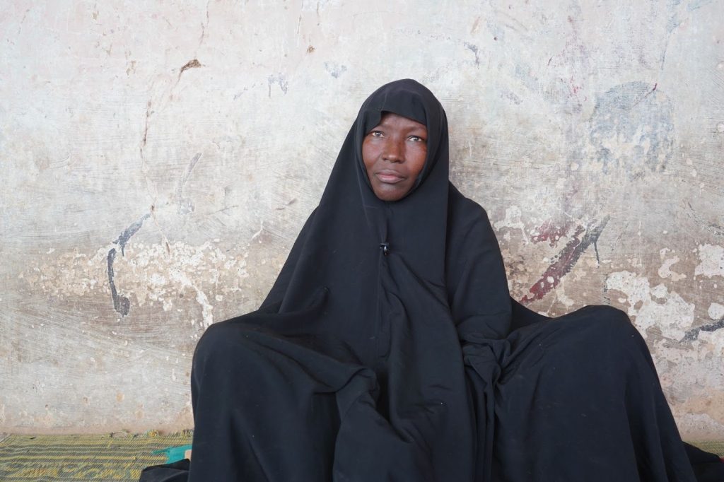 Person in a black cloak sits against a textured, weathered wall, gazing thoughtfully upwards.