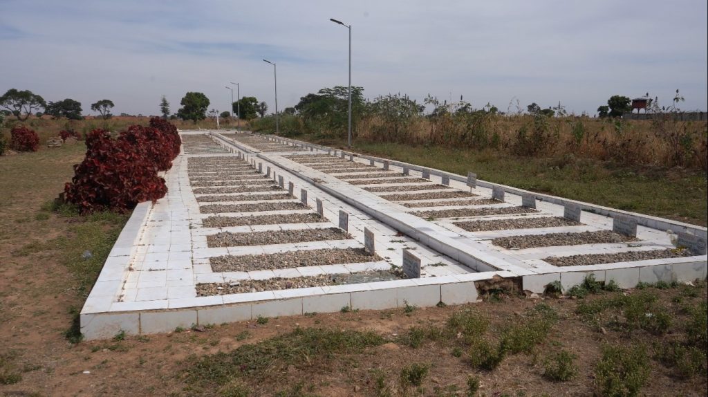 A series of rectangular plots with gravel sections, lined with small plaques, bordered by grass and trees under a cloudy sky.