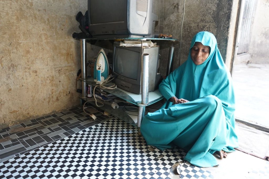 Person in a turquoise outfit sits near a shelf with old electronics and an iron, on a checkered floor in a dimly lit room.