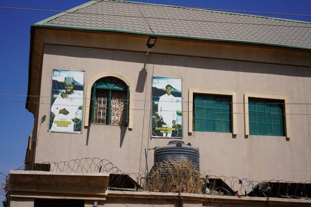 A beige building with green windows, barbed wire barrier, and two memorial posters featuring men in white attire with the text "Our Immortal."