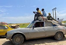 People sit atop a heavily loaded car on a rural road, with fields and power lines in the background under a clear blue sky.