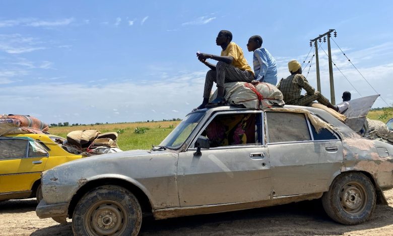 People sit atop a heavily loaded car on a rural road, with fields and power lines in the background under a clear blue sky.