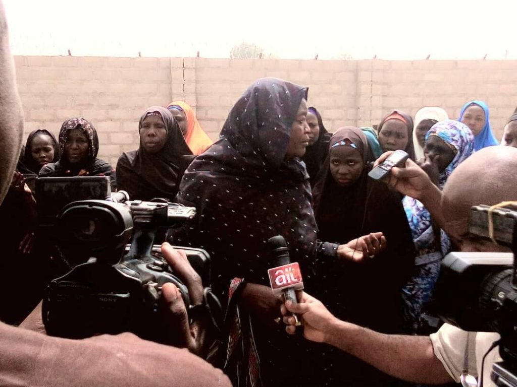 A woman speaks to the press, surrounded by a group of women in colorful headscarves, with microphones and cameras capturing the scene.