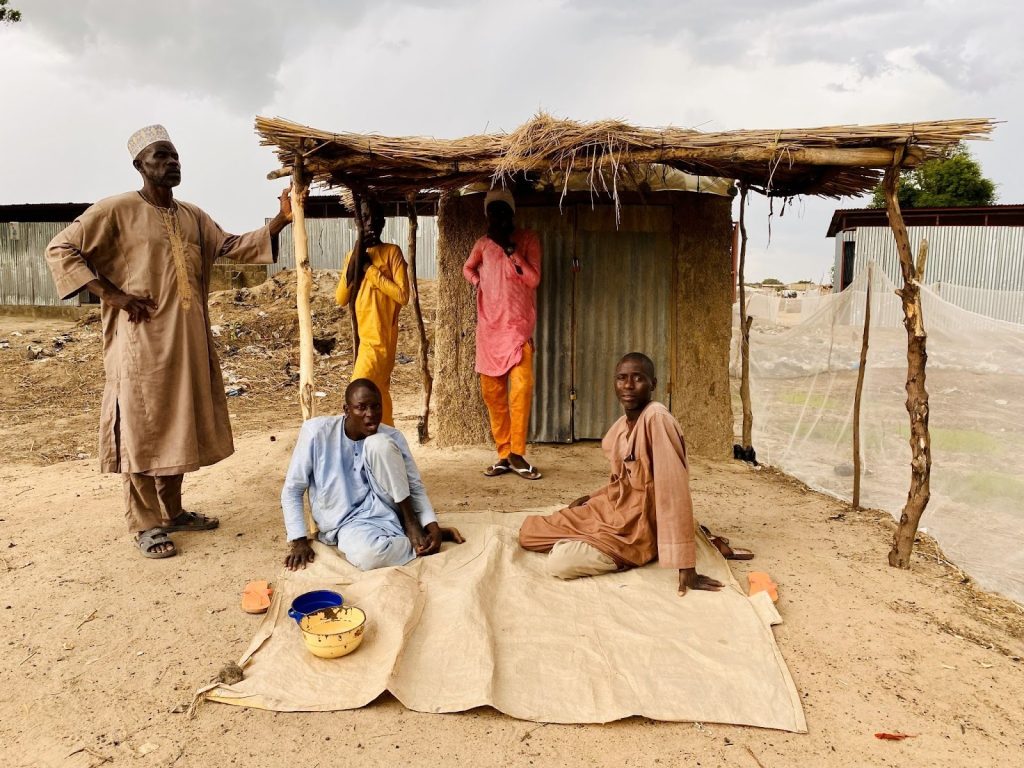 Four men in traditional clothing under a makeshift shelter in a rural area, with bowls on a mat in the foreground.