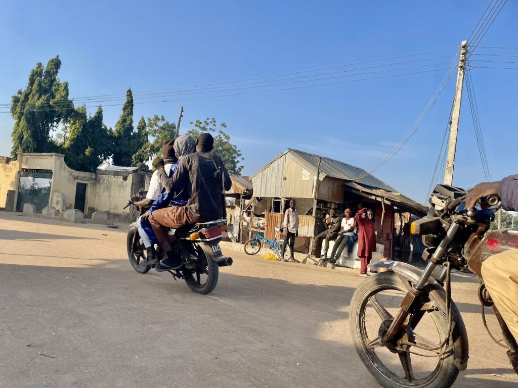 Three people ride a motorcycle on a sunny street, with bystanders and old buildings in the background.