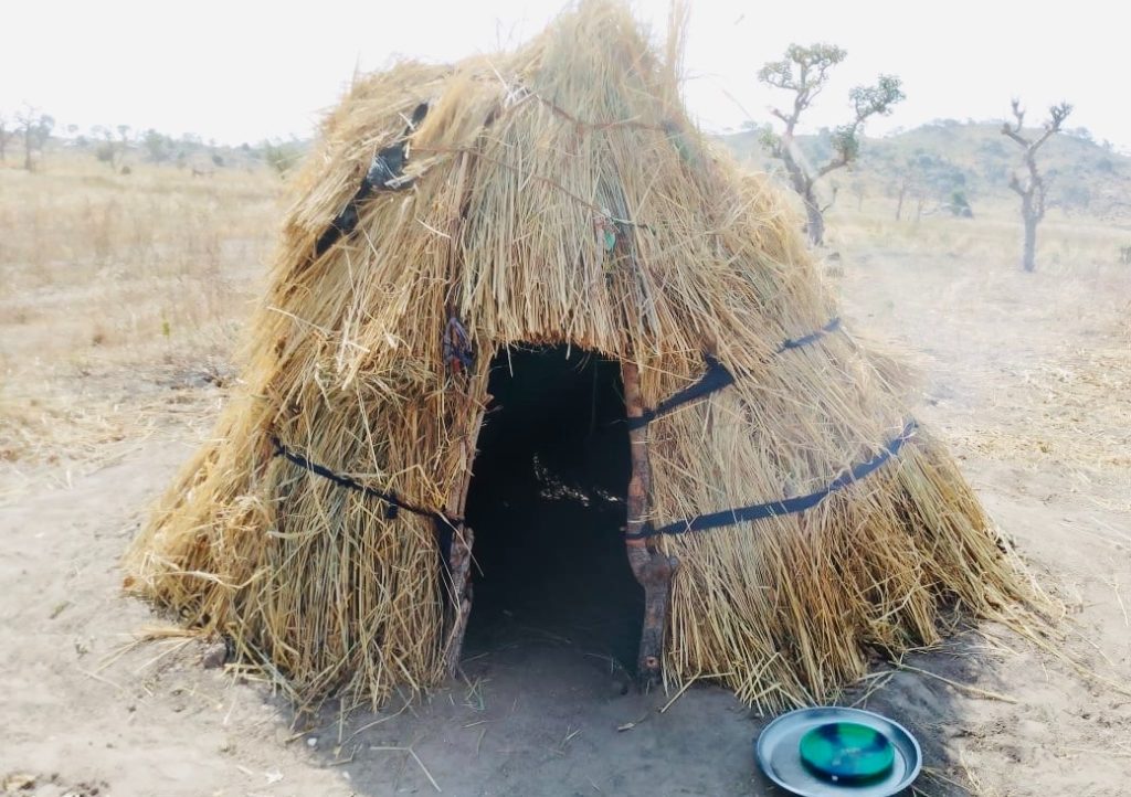 Small, round hut made of straw and twigs with a narrow entrance. It stands on dry ground with sparse trees in the background.