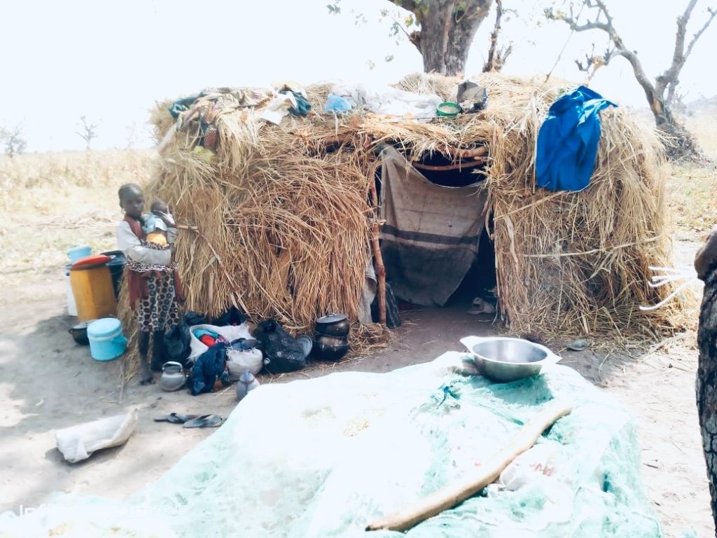 A child stands outside a small straw hut with belongings scattered nearby in a rural area, with trees and dry grass in the background.