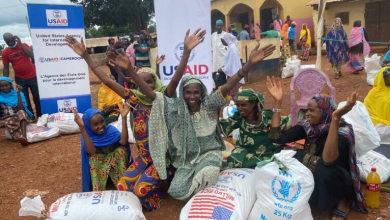 Group of people celebrating near aid supplies, with USAID banners visible in the background, in an outdoor setting.