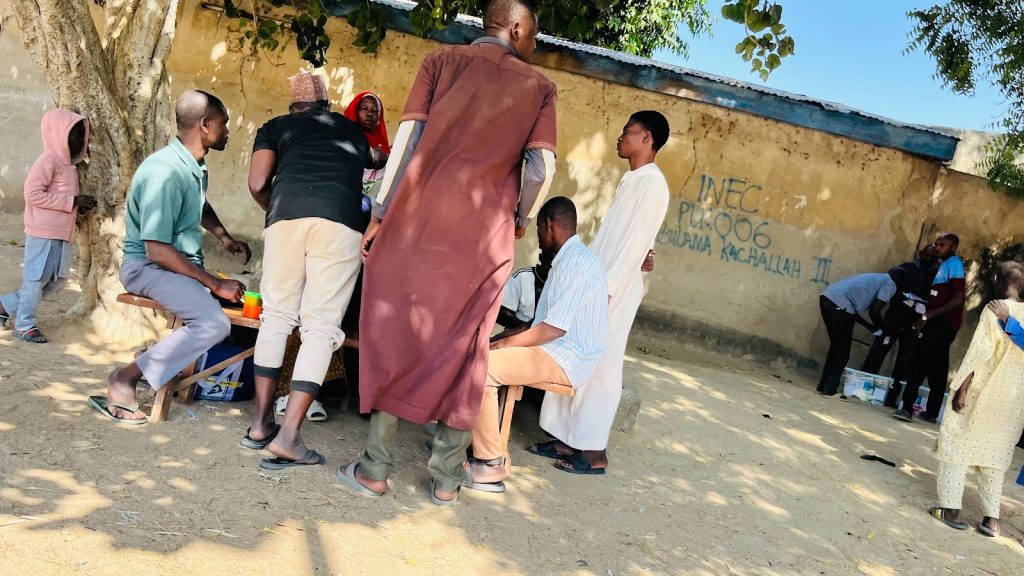 People gather around a table outdoors, near a wall with writing. Trees provide shade in the background.