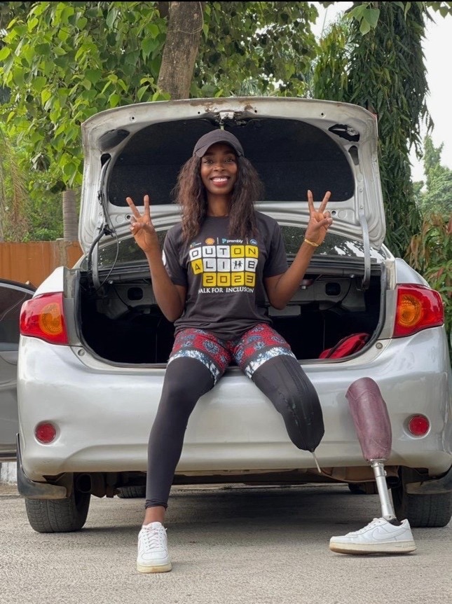 Smiling woman with a prosthetic leg sits in open car trunk, wearing a cap and "Out On A Limb 2023" shirt, showing peace signs.