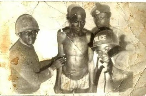 A shirtless man is flanked by three uniformed officers with helmets, one labeled "POLICE," in a grainy, aged black and white photo.