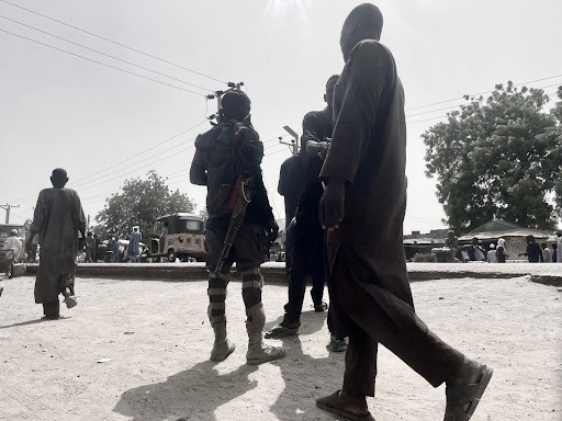A group of people, including a person equipped with gear, walking in a sunlit area with trees and vehicles in the background.