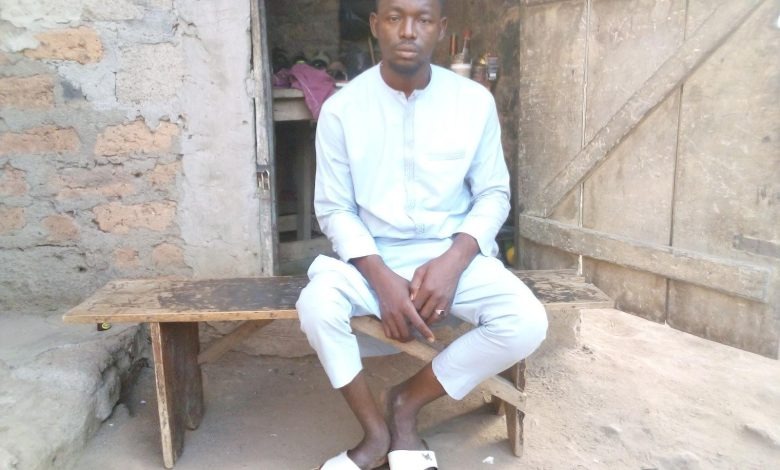Man in light blue traditional attire sits on a wooden bench in front of a rustic wall, with a doorway visible behind him.