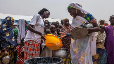 Women and children gather to receive food from a volunteer with a yellow bucket at a distribution event.