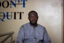 A man in a gray shirt sits with a calm expression in front of a wall with "DON'T QUIT" written behind him.