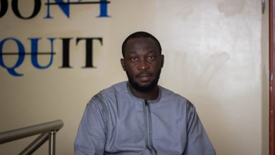 A man in a gray shirt sits with a calm expression in front of a wall with "DON'T QUIT" written behind him.