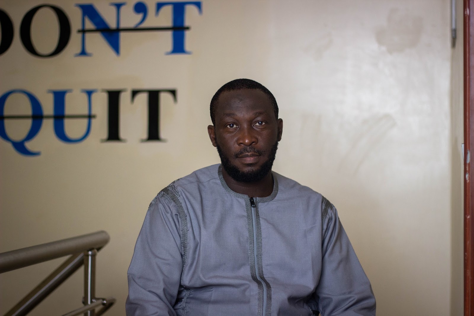 A man in a gray shirt sits with a calm expression in front of a wall with "DON'T QUIT" written behind him.
