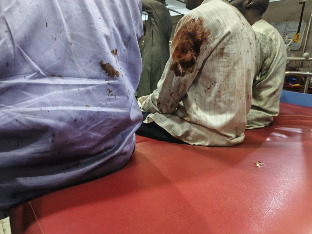 Three individuals sit on a red mat with stained shirts, showing signs of wear, in a room with medical equipment.